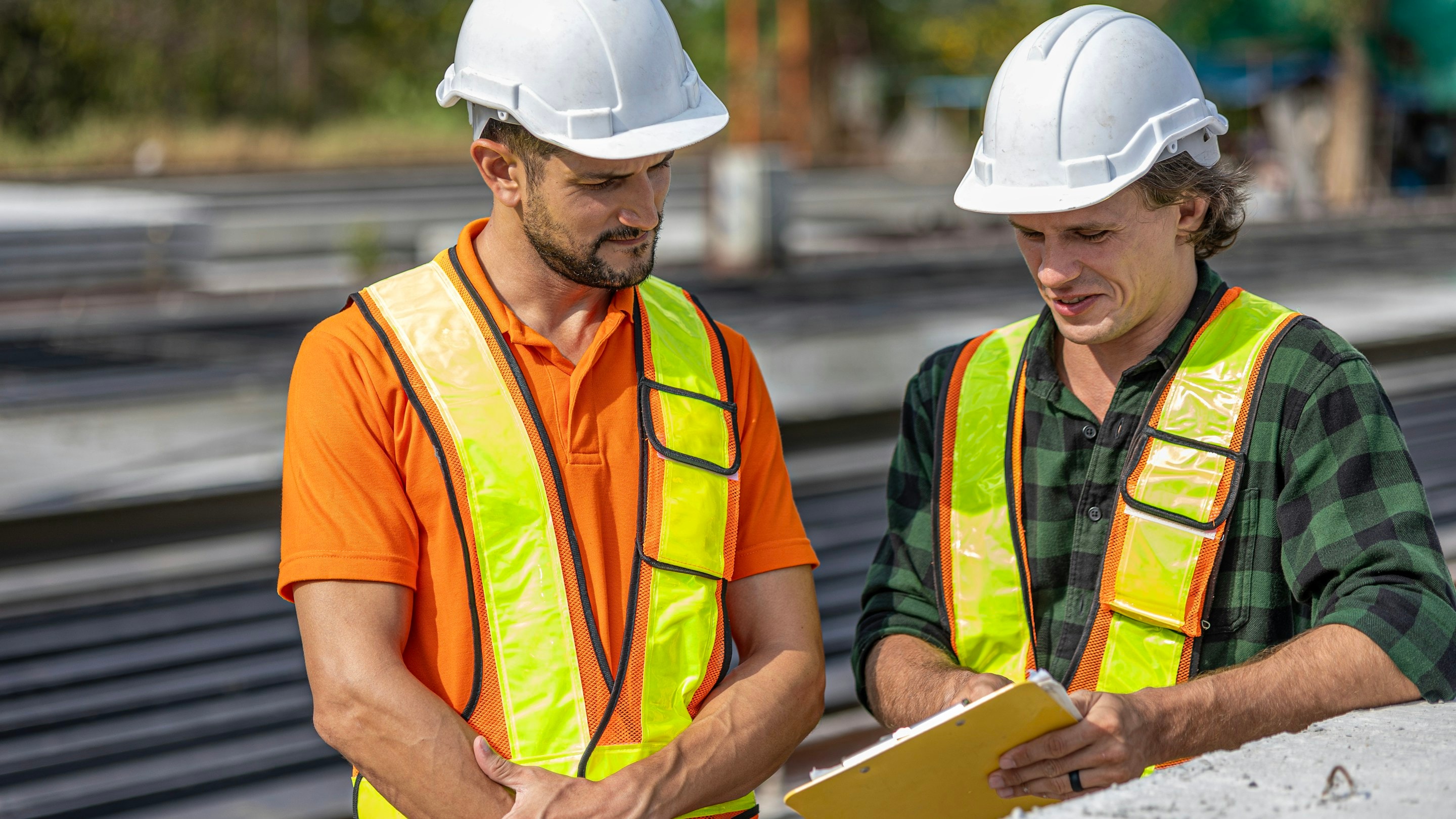 Two construction workers in hard hats and safety vests reviewing plans on a clipboard