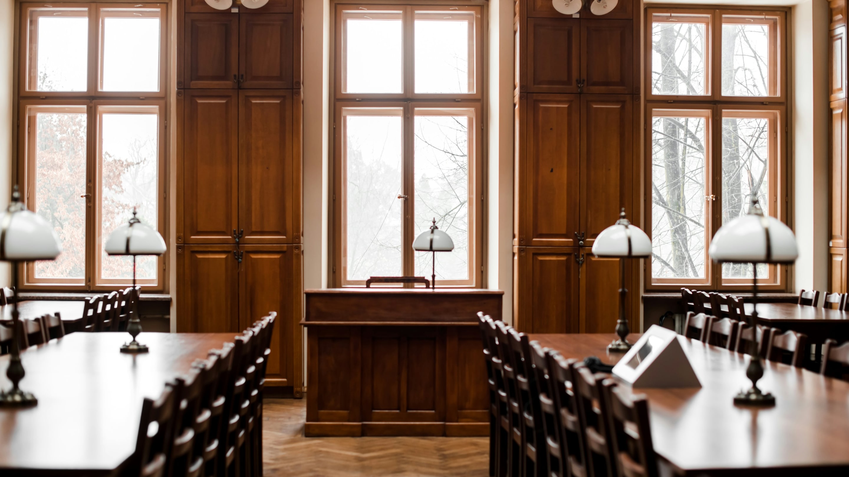 A wood-paneled courtroom with tall windows and brass desk lamps on the counsel tables